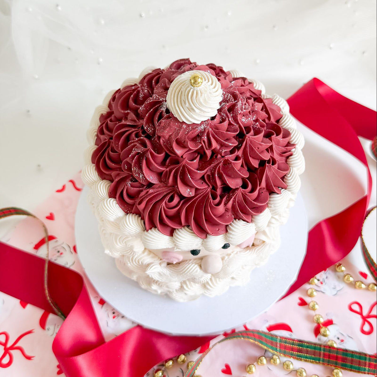 A decorated cake shaped like Santa's face, topped with dark red piped frosting resembling Santa's hat and white piped frosting resembling his beard, set on a white plate with red ribbons and holiday decorations around it.