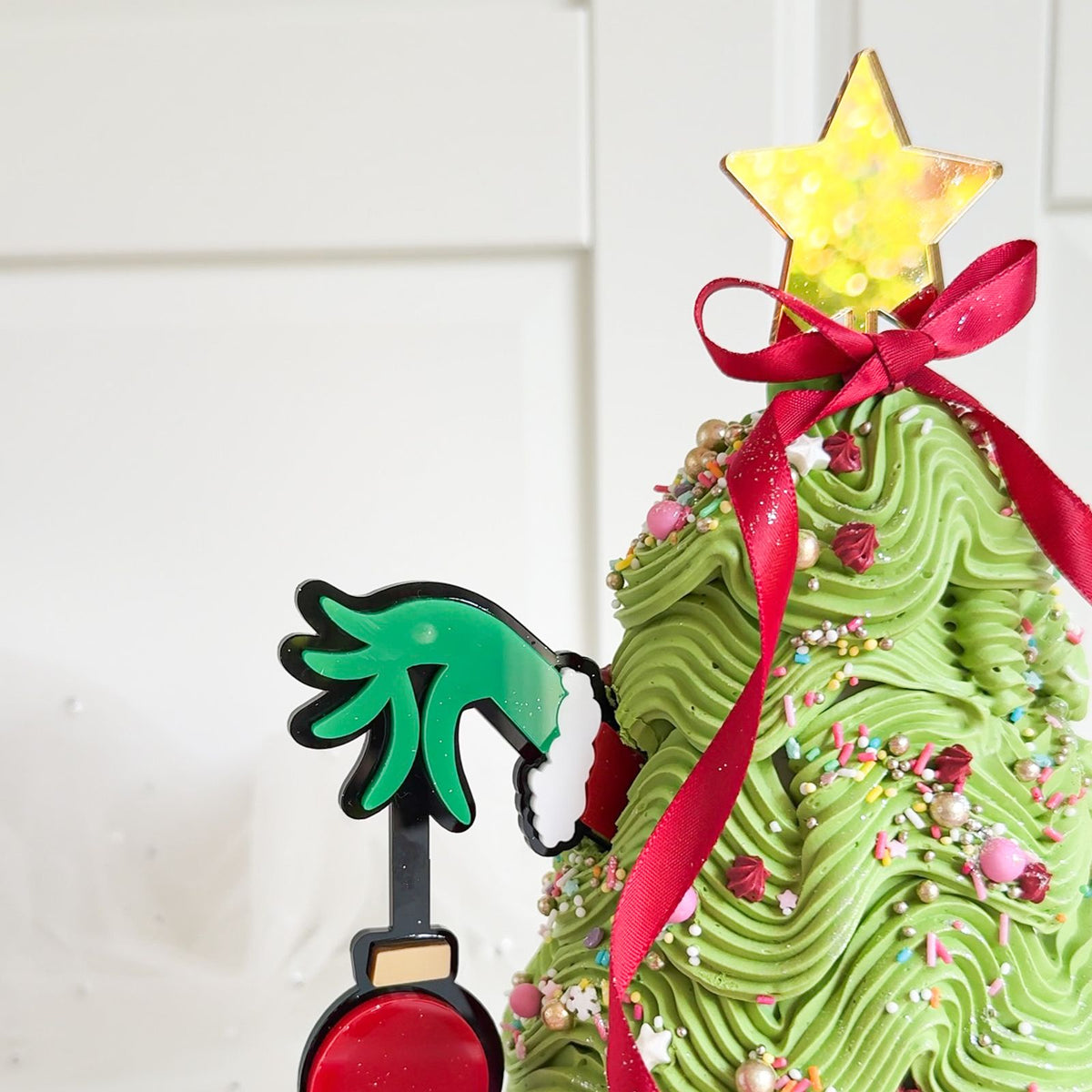 A close-up of a decorated green iced cake styled like a Christmas tree, topped with a gold star, red ribbon, and sprinkles, with a Grinch-themed topper on the left against a white paneled background.