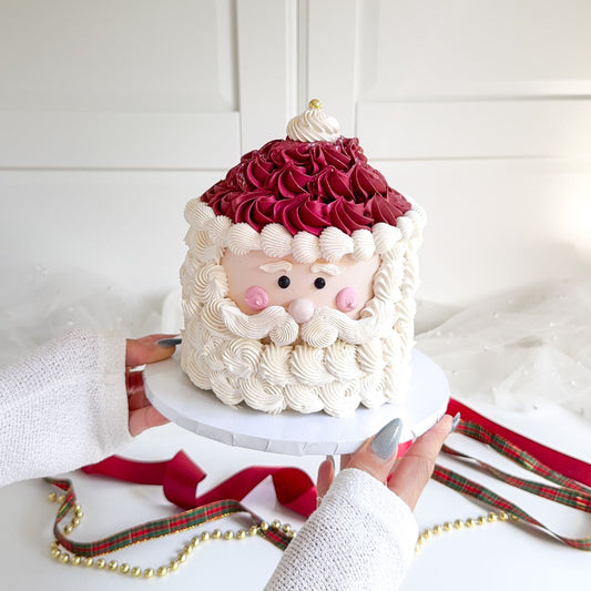 A Santa Claus themed cake decorated with cream-colored piped frosting beard and face, and a deep burgundy frosting hat, held on a white cake stand by hands wearing white knit sleeves.