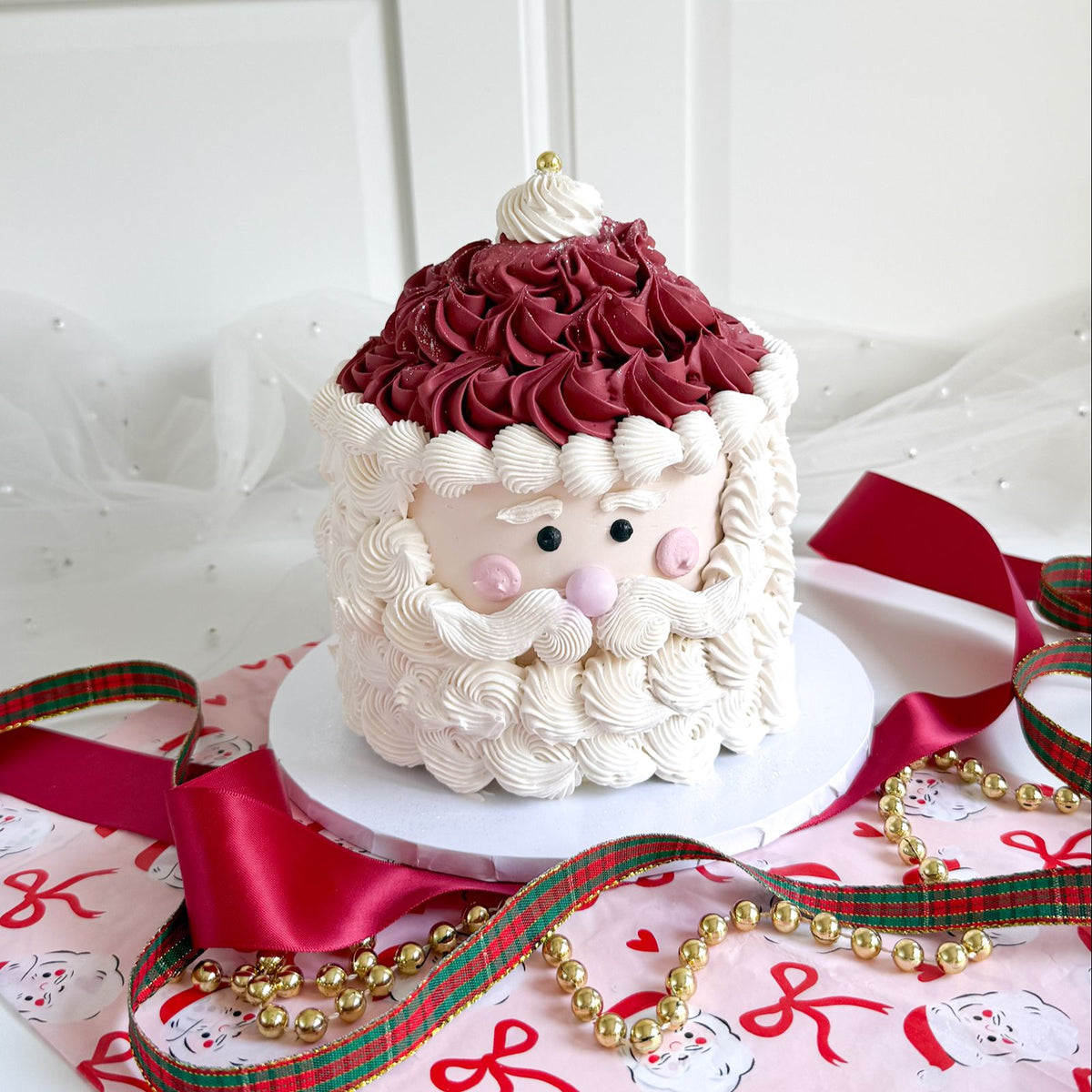 A Santa Claus-themed birthday cake decorated with piped white frosting for the beard and face, dark red frosting for the hat, and small pink cheeks, displayed on a white cake stand with red ribbons and gold beads.