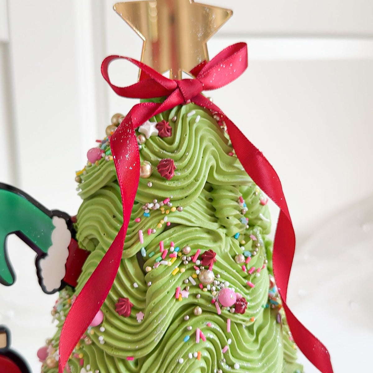 A close-up of a decorated green frosting Christmas tree with a gold star topper, red ribbon bow, and colorful sprinkles, set against a bright white background.